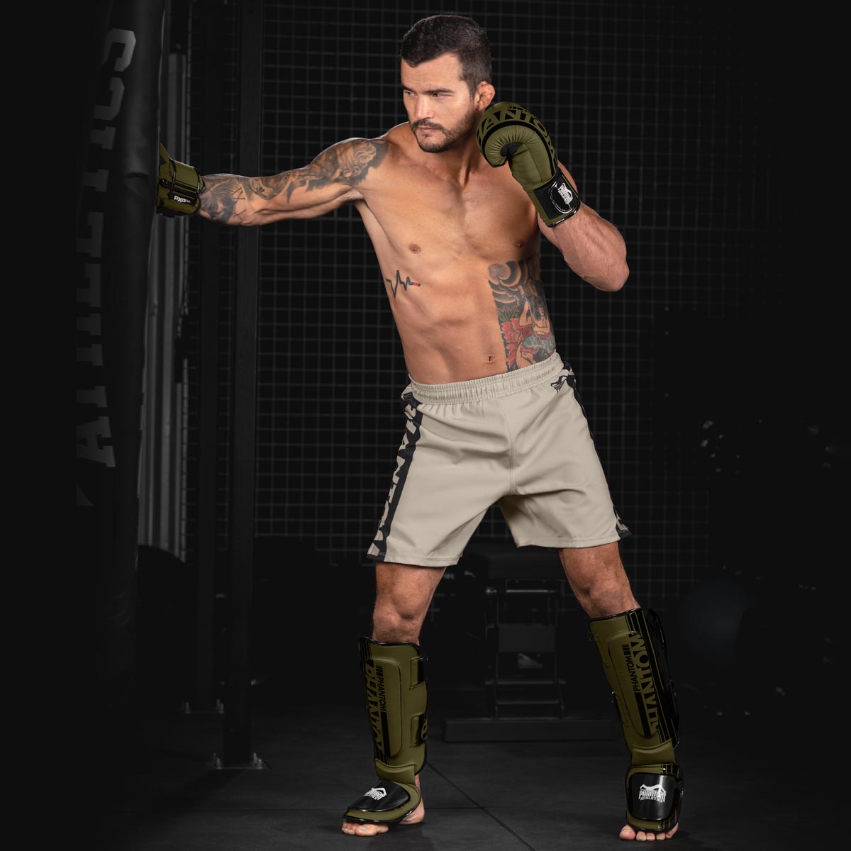 MMA Fighter beim Training am Boxsack mit dem Phantom Apex Fightequipment in Army Grün. Bestehend aus Phantom Apex Boxhandschuhen, MMA Shorts und Hybrid Schienbeinschonern.